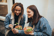 © polinaloves - Two young women female friends using smartphone sitting outdoors eating takeaway food, laughing and having fun. Food delivery and takeout.