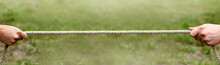 Two Hands Hold A Long Rope Against The Background Of A Green Lawn. Outdoor Tug Of War