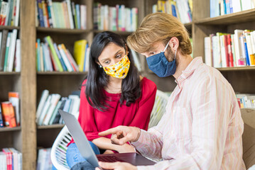 Wall Mural - young couple of multiracial students using laptop in a library during Corona Virus epidemic, wearing face mask fro protection, team work and health care concepts