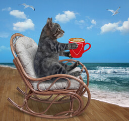 Wall Mural - A gray cat with a cup of coffee is in a wooden rocking chair by the sea.