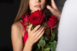© LIGHTFIELD STUDIOS - cropped view of blurred woman holding red roses near man on dark background.