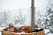 Woman relaxing in hot bath outdoors, enjoying thermal spa at snowy mountains. Winter holidays in the mountains, hot water treatments concept. Caucasian woman wearing winter hat