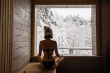 Young woman sitting in sauna with beautiful view on snowy forest. Concept of winter SPA and recreation on nature. Idea of scandinavian recreation and lifestyle