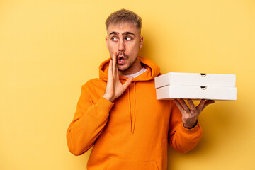 Wall Mural - Young caucasian man holding pizzas isolated on yellow background is saying a secret hot braking news and looking aside