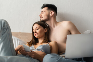 Wall Mural - shirtless man and sexy woman in bra and jeans holding gadgets while relaxing with closed eyes in bedroom.