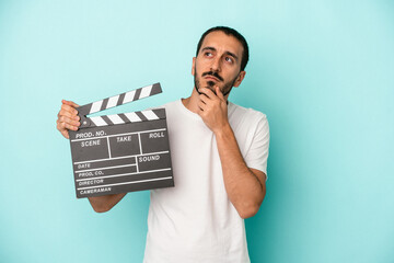 Young caucasian actor man holding clapperboard isolated on blue background looking sideways with doubtful and skeptical expression.