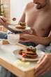 © LIGHTFIELD STUDIOS - partial view of smiling shirtless man eating tasty pancakes with strawberries near blurred girlfriend in bedroom.