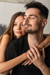 Wall Mural - happy man with closed eyes touching hands of tender woman hugging him in bedroom.