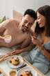 © LIGHTFIELD STUDIOS - cheerful woman in jeans and bra feeding boyfriend with strawberries during breakfast in bedroom.