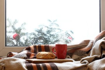 Wall Mural - plaid, a red mug and pastries on a wooden stand against the background of a window in winter. warm cozy home