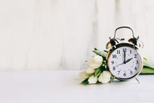 Daylight Savings Time Concept. Set Your Clocks And To 2 Am And Spring Ahead With This Image Of An Alarm Clock With White Tulip Flowers. Selective Focus With Blurred Foreground And Background With Copy