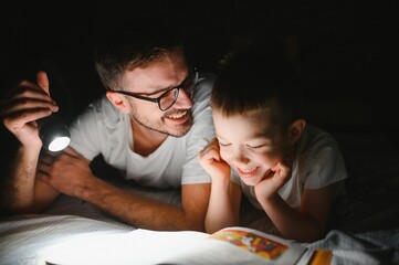 Happy family reading bedtime story under blanket in evening. Father and son spend time together. Father's Day.