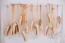 Many Hanging Ballet Shoes On White Wall Background In Studio. New Pointe Shoes With Satin Ribbons Hanging On Rank. Ballet Shoes Hang On Bar In Room. Concept Of Dance, Ballet School, Ballerinas Clothes