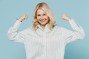 Wall Mural - Elderly strong sporty woman 50s wear striped shirt showing biceps muscles on hand demonstrating strength power isolated on plain pastel light blue background studio portrait. People lifestyle concept.