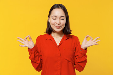 Spiritual tranquil young woman of Asian ethnicity 20s wear orange shirt hold spreading hands in yoga om aum gesture relax meditate try to calm down isolated on plain yellow background studio portrait