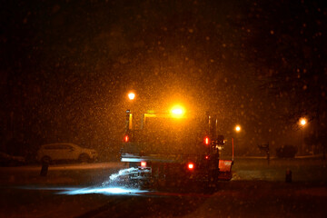 SEWICKLEY, PA, USA - JANUARY 17TH 2022: a vehicle is spraying salt on a snow-covered residential street lit by street lights late at night. The snow’s 8 inches deep and the road is impassable for cars