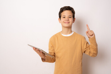 Cute boy in casual t-shirt holding tablet pointing finger up over white background.