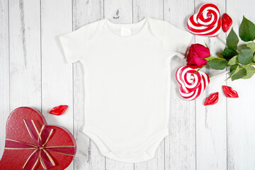 Baby romper onesie jumpsuit product mockup. Valentine's Day farmhouse theme SVG craft product mockup styled with red roses, and heart shaped lollipops against a white wood background. Flatlay.