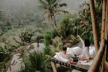 Young Travelling Couple Relaxing In The Jungle Resort Hotel In Bali, Indonesia Surrounded By Rice Fields, Palm Trees And Lush Green Landscape