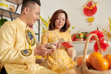 Poster - Positive couple in traditional dresses putting money in red envelopes for Chinese New Year celebration