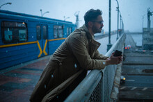 A Bearded Man With Glasses In A Winter Coat, Standing Leaning On The Railing Of A Bridge Over A Canal, Deep In Thought, Looking Straight Ahead.