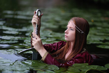 Red Haired Woman In Red Medieval Dress Holding Long Sword Stands In Lake With Lily Pads. Fantasy Portrait Of Lady Warrior