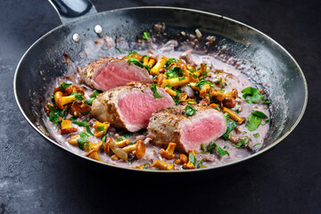 Fried dry aged pork fillet chateaubriand medallion steak natural with chanterelles in walnut cream sauce served as close-up in a classic skillet on black background