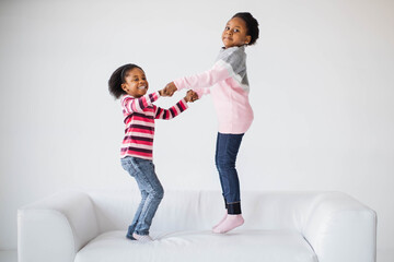 Wall Mural - Pretty african american children holding hands and jumping together on white cozy sofa. Two happy sisters with curly hair enjoying carefree lifestyles during childhood.