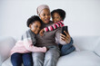 © sofiko14 - African american woman sitting with two pretty daughter in couch and taking selfie on modern smartphone. White studio background. Family and gadgets concept.