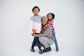 Poster - Cheerful african american islamic family of three smiling and hugging over white background. Caring mother and her two little daughters spending time with fun indoors.