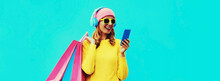 Colorful Portrait Of Stylish Smiling Young Woman Listening To Music In Headphones With Phone And Shopping Bags Wearing Yellow Knitted Sweater, Pink Hat On Blue Background