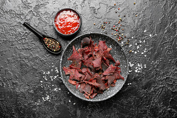 Plate with spicy beef jerky and sauce on dark background