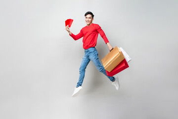 Wall Mural - Surprised Asian man holding bags and red envelope Ang Pow   jumping in isolated light gray studio background for Chinese new year shopping concept