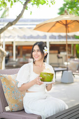 Wall Mural - Beautiful woman tourist with white flower on her hair drinking coconut sitting on lounge chair during summer holidays