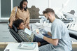 © pressmaster - Contemporary stomatologist showing false teeth to little African patient sitting in armchair in front of him in dental clinics