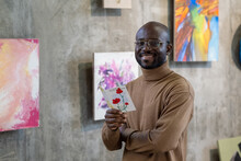 Cheerful Young African Man With Leaflet Looking At You While Standing Against Wall With Paintings In Art Gallery