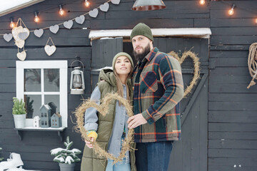 Wall Mural - young couple in love beautiful Asian girl and bearded man in stylish winter clothes standing near porch of country house with handmade wreath of dried grass in shape heart, winter spirit valentine day