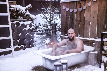 Wall Mural - young brutal bearded tattoo man sit in bath with hot water on the snow near porch of country house with wooden fence decorated to celebrate Valentine's Day in suburbs