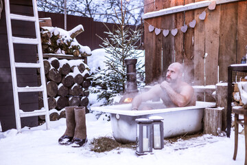 Wall Mural - young brutal bearded tattoo man sit in bath with hot water on the snow near porch of country house with wooden fence decorated to celebrate Valentine's Day in suburbs