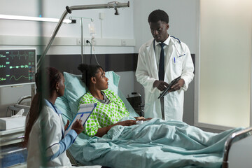 Wall Mural - African american therapist doctor checking sick woman holding tablet with medical expertise discussing healthcare treatment. Patient resting in bed during clinical consultation in hospital ward