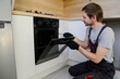 © glebcallfives - Young man worker in uniform repairing electric oven in kitchen.