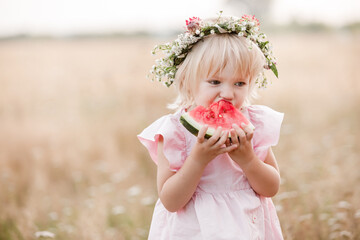 Wall Mural - picnic with family. cute little girl eating big piece of watermelon on the grass in summertime in the park. Adorable child wearing in flowers wreath on head and pink dress.