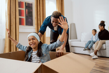 Happy funny African American family moving into new apartment, little preschool daughter sitting in boxes, father rolling her, real estate purchase concept