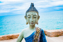 Sculpture Of A Meditating Yogi In White Clay And Blue Tones With The Face Of Buddha With The Relaxing Sea In The Background Out Of Focus.