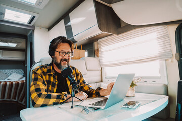 Young adult use microphone and laptop computer to podcast web content inside a camper van. Digital nomad lifestyle people. Recording creative job. Male working with online roaming connection