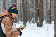 Young Man In Knitted Hat With Backpack Looking At Map On Smartphone While Hiking In Winter Forest Using Modern Technology Navigation