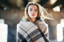 Close-up Portrait Of Beautiful Young Woman Wrapped In Wool Blanket Looking To Camera On Windy Sunny Day On Urban Street Background