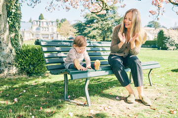 Wall Mural - Outdoor portrait of happy young mother playing with adorable baby girl in spring park, sitting on the bench, clapping with hands