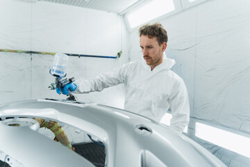 Wall Mural - Young curly car painter varnishes a car bumper. Man works in a spray booth. Process of painting car body parts