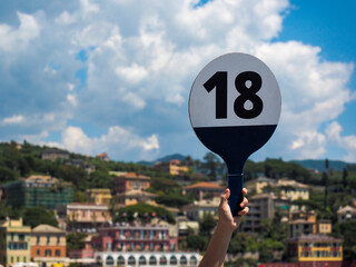 Tour Guide holding board with tour Number on with defocused Mediterranean village in the background